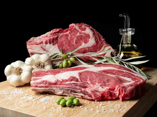 Dos chuletones Vuelta y Vuelta sobre tabla de madera, con sal esparcida, un ajo, una aceitera con AOVE y una rama de olivo con aceitunas verdes