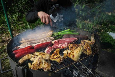 Un hombre con un delantal de Vuelta y Vuelta está manipulando una barbacoa humeante con distintos tipos de carne de ternera, morcilla, panceta, chorizos, espárragos y alitas de pollo