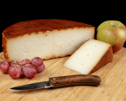 Queso Majorero en tabla, medio queso y delante una cuña. Aparece también una navaja, uvas y una manzana