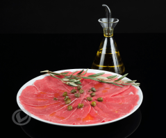 Carpaccio de ternera Vuelta y Vuelta, en finas lonchas en un plato con alcaparras por encima. También se ve una rama de olivo y una aceitera con AOVE