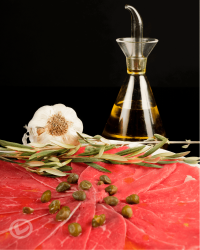 Carpaccio de ternera Vuelta y Vuelta, en finas lonchas en un plato con alcaparras por encima. También se ve un ajo, una rama de olivo y una aceitera con AOVE