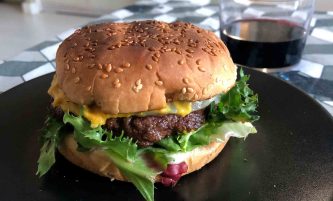 Hamburguesa de ternera ya montada en pan, con su lechuga, tomate, cebolla y al fondo, un vaso de vino