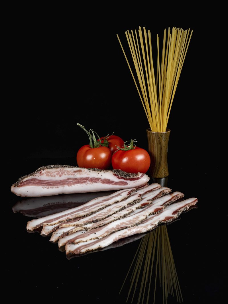 Trozo de guanciale y varias lonchas cortadas delante. También tres tomates y un botecito con spaguettis