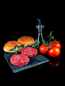 Hamburguesas de carne picada de terneras Vuelta y Vuelta sobre una pizarra. Detrás dos panes de hamburguesa. También tomates